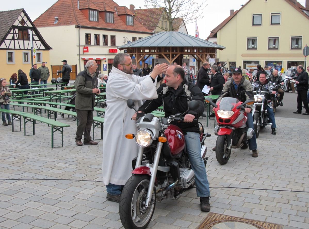 Auf einem Marktplatz sitzt ein Mann auf einem Motorrad. Neben ihm steht ein Mann mit einem weißen Gewand und segnet den Motorradfahrer.
