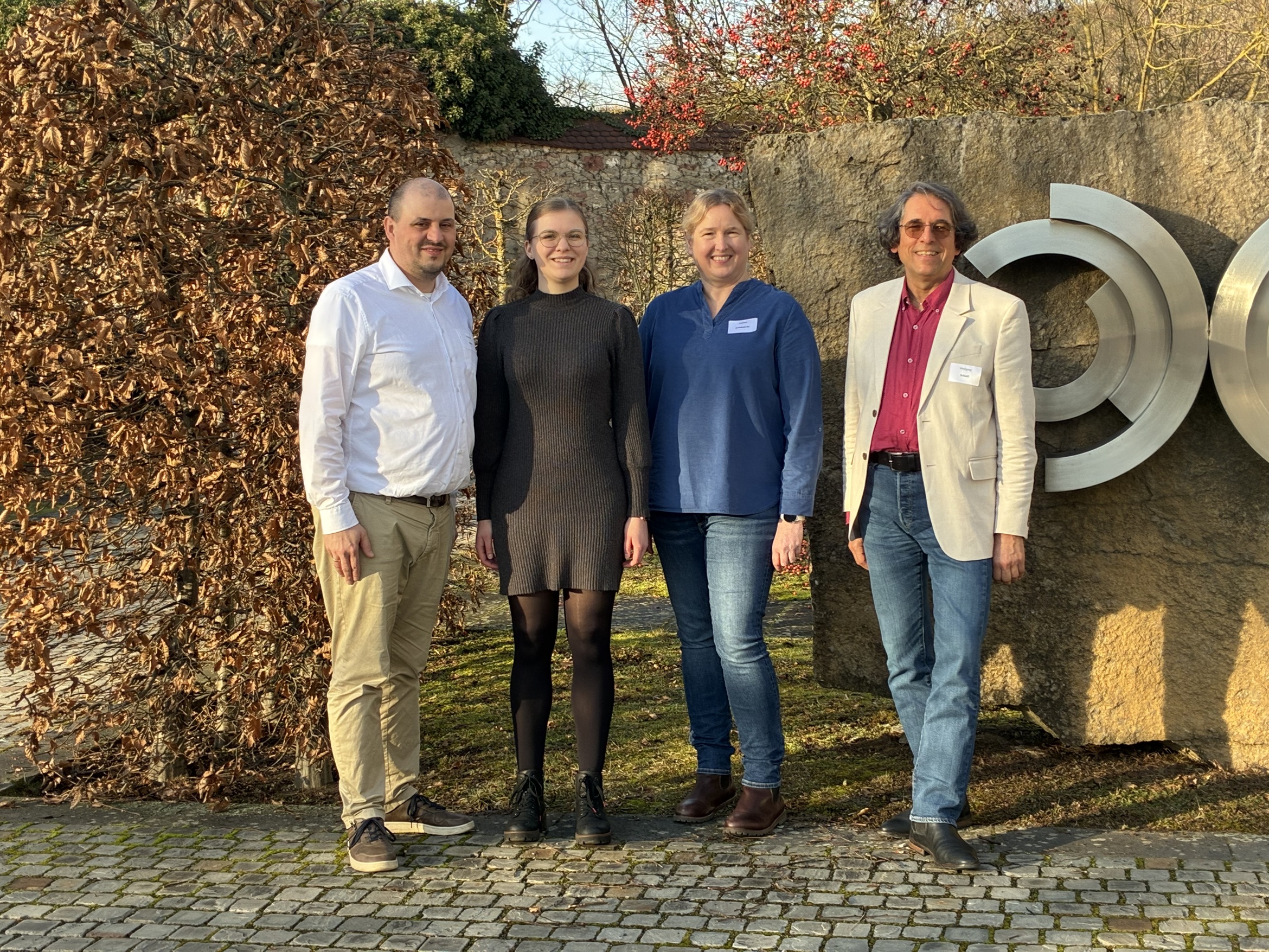 Ein Gruppenbild mit vier Personen, die nebeneinander stehen. Im Hintergrund ist eine Hecke und eine Natursteinwand zu sehen.