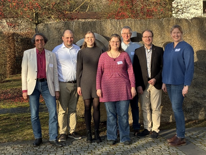 Ein Gruppenbild mit sieben Personen. Im Hintergrund sind man den Teil eines Hauses, Bäume ohne Blätter und einen blauen Himmel.