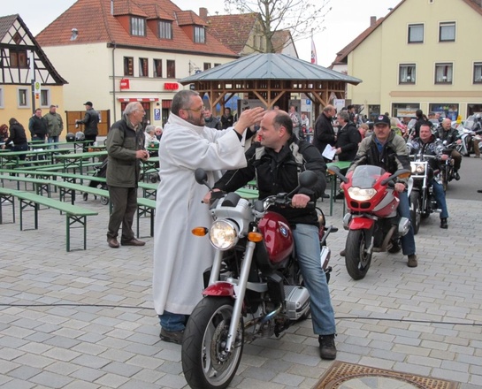 Auf einem Marktplatz sitzt ein Mann auf einem Motorrad. Neben ihm steht ein Mann mit einem weißen Gewand und segnet den Motorradfahrer.
